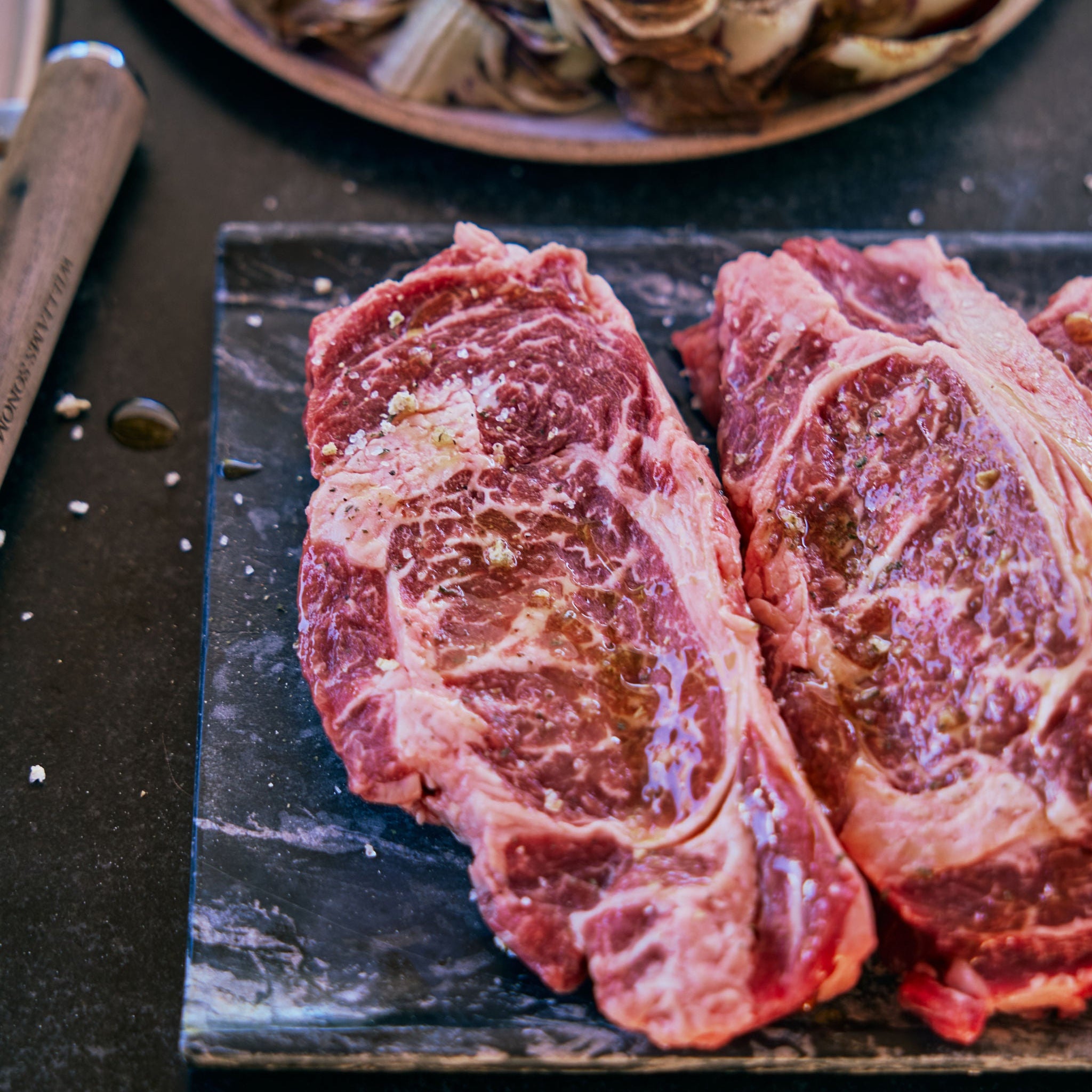 Two raw ribeye steaks on a black slate board with artichokes in the background.