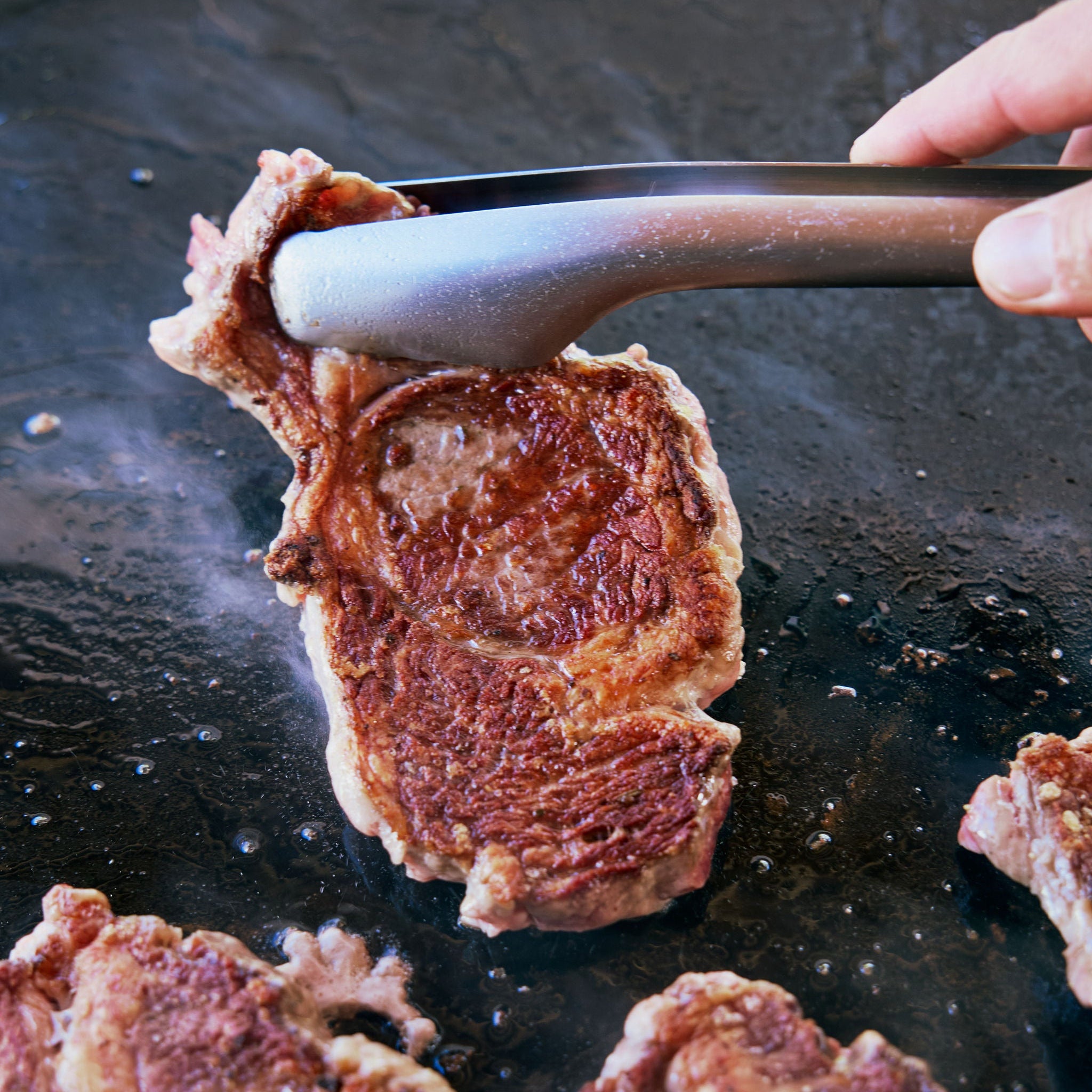 Person using tongs to flip a piece of seared meat on a hot surface