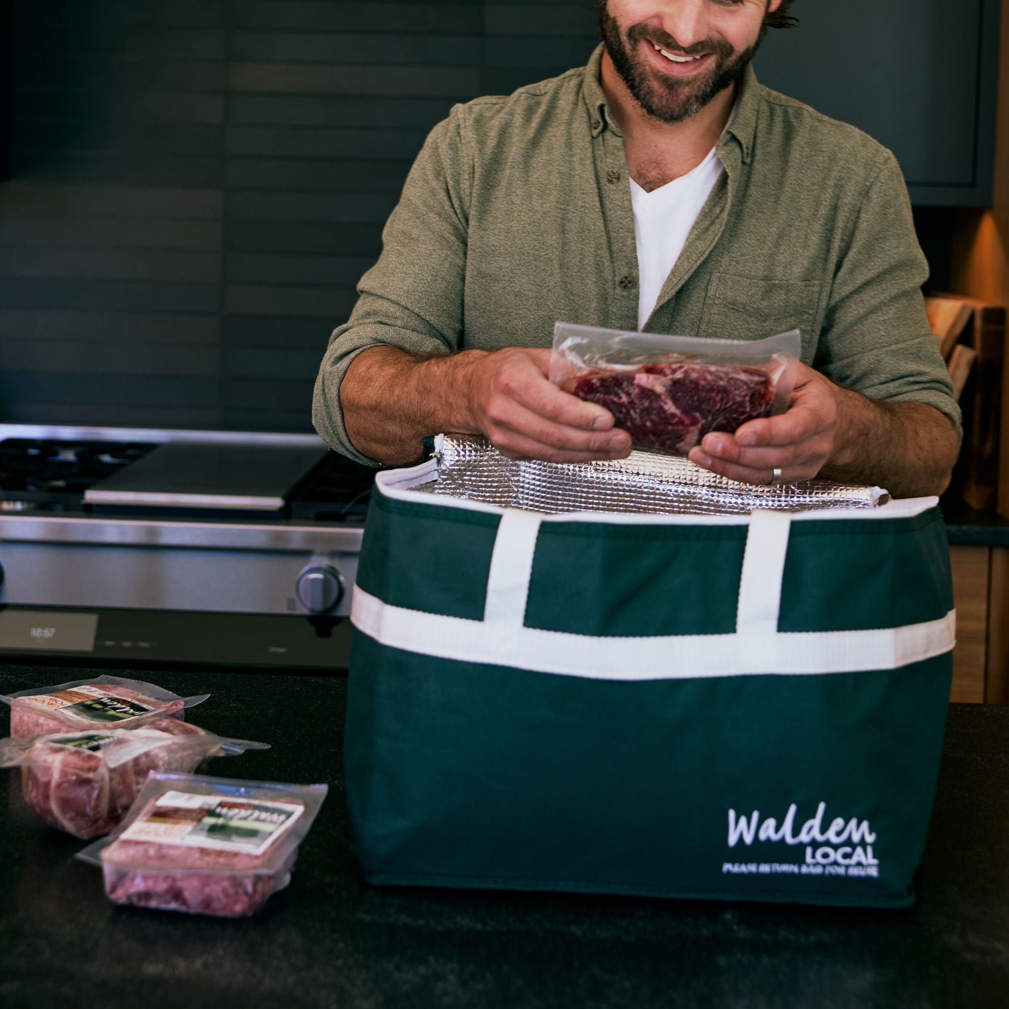 Man in a kitchen holding a package of meat next to a Walden Local cooler bag.